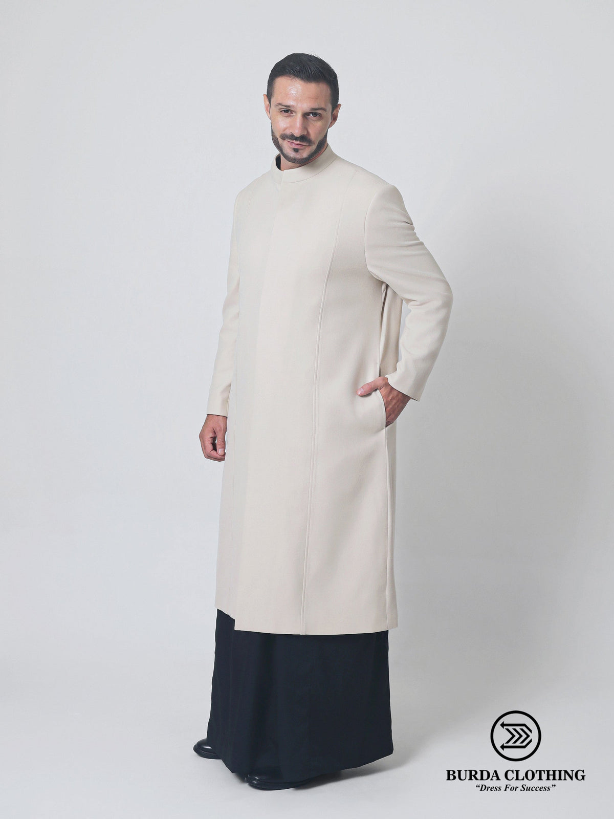 A man modeling a long-sleeved, high-neck cream-colored coat over a dark navy thobe, standing against a light gray studio background. The Burda Clothing brand logo is visible in the bottom right corner.